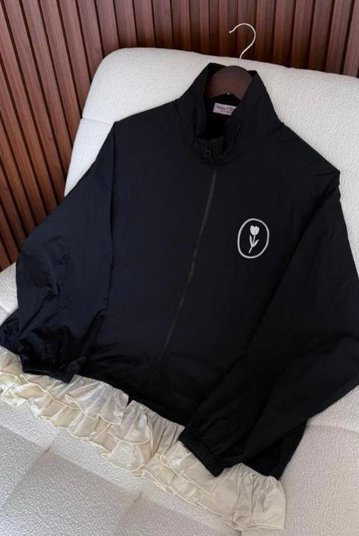 A black lightweight zip-up jacket by Haidee Orlin with a small white circular flower logo on the left chest, laid flat on a white textured chair in front of a brown slatted wall.