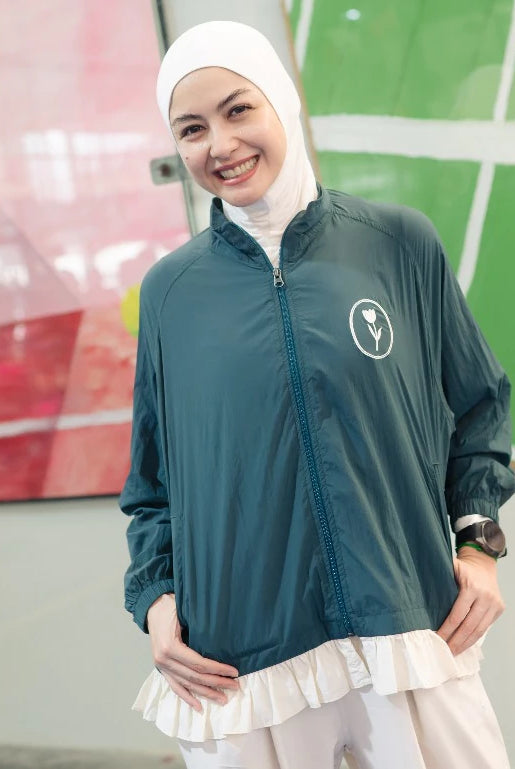 A woman wearing a dark teal green lightweight zip-up jacket by Haidee Orlin with a white ruffled hem, a white hijab, and white pants, standing in front of a modern wall with abstract art.