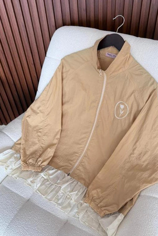 A beige lightweight zip-up jacket by Haidee Orlin with a small white circular logo on the chest, draped over a white textured chair in front of a brown slatted wall.