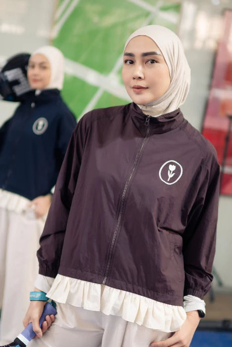 A woman wearing a white hijab and a dark plum zip-up jacket by Haidee Orlin with a white ruffled hem stands in an indoor sports facility. Another person is blurred in the background holding a camera.