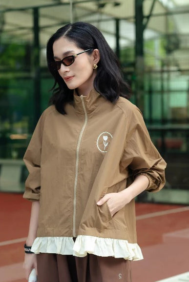 A woman wearing a brown lightweight zip-up jacket by Haidee Orlin with a white ruffled hem and a small circular chest graphic, standing in an outdoor modern architectural setting.