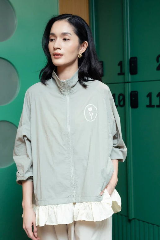 A woman wearing a light sage green zip-up jacket by Haidee Orlin with a white circular logo on the chest, layered over a cream ruffled top and cream pants, standing in front of green metal lockers.