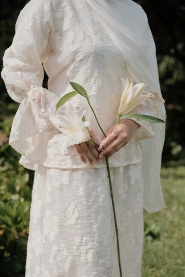 Person wearing a broken white lace blouse with floral embroidery by Renaette holding a flower in a natural setting