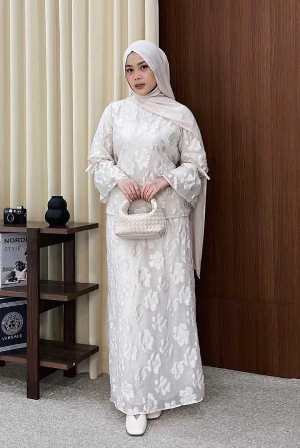 Woman in a white floral outfit by Renaette standing in a room with a wooden side table and books.
