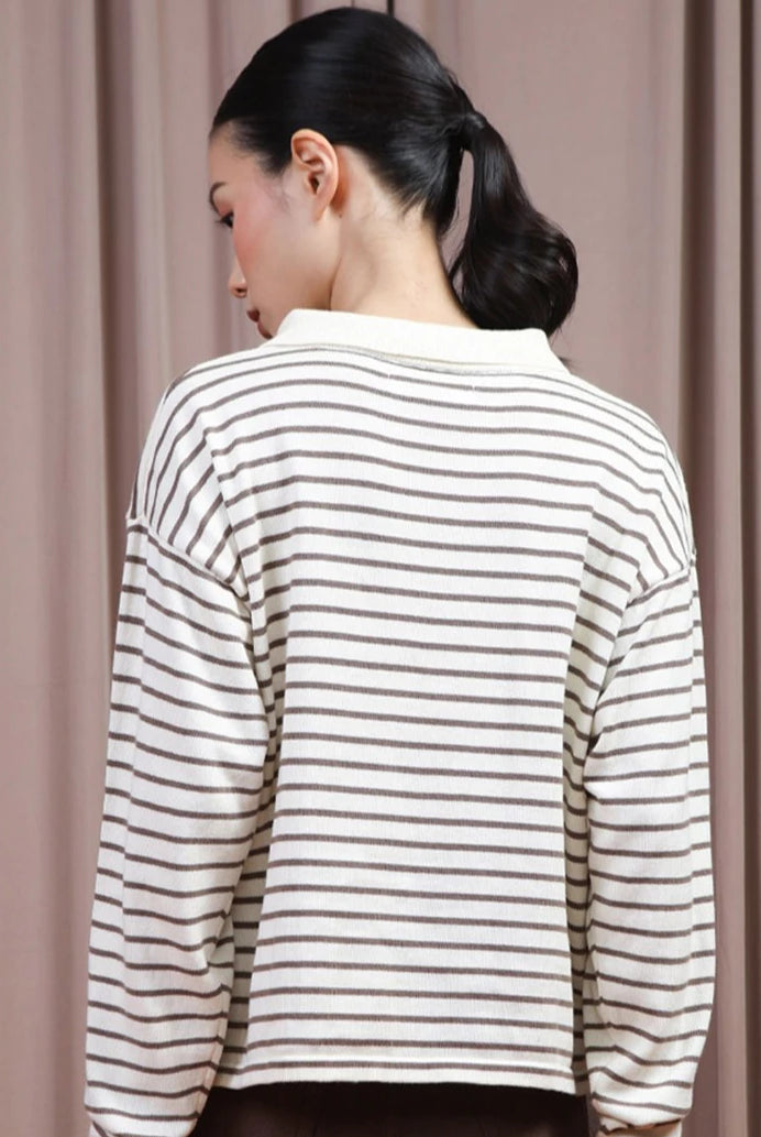 A woman shown from the back wearing a cream-colored long-sleeve polo shirt by Hijabchic with thin brown horizontal stripes, set against a soft beige curtain background.