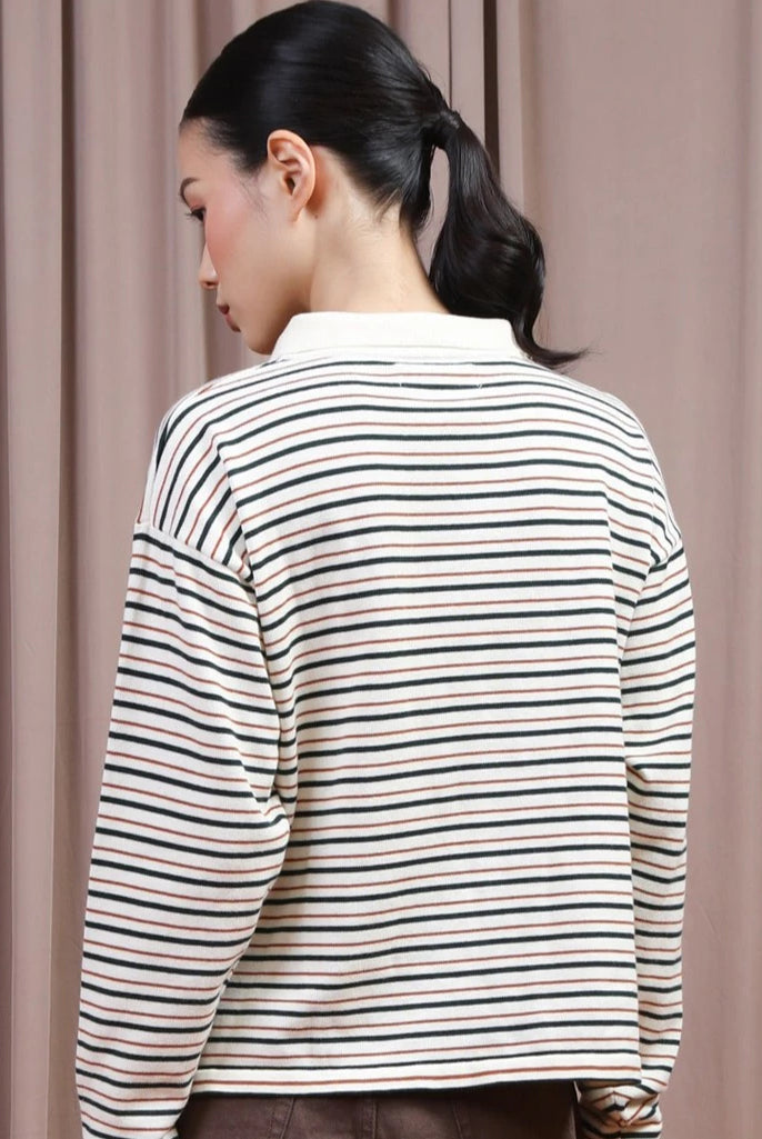 A woman shown from the back wearing a long-sleeve striped polo shirt by Hijabchic with thin horizontal green, black, and beige stripes on an off-white base, set against a soft beige curtain background.