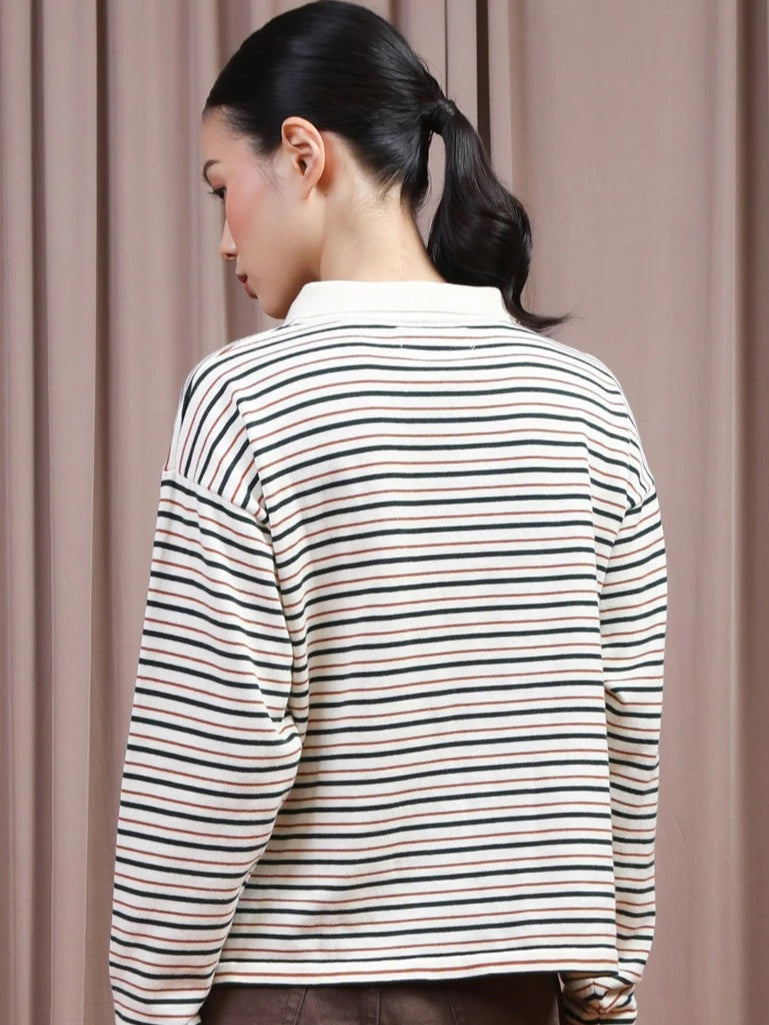 A woman shown from the back wearing a long-sleeve striped polo shirt by Hijabchic with thin horizontal green, black, and beige stripes on an off-white base, set against a soft beige curtain background.