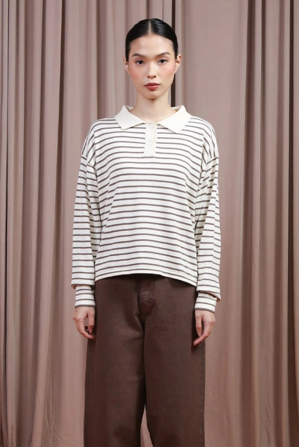 Person wearing a brown and white striped shirt by Hijabchic and brown pants against a beige curtain background