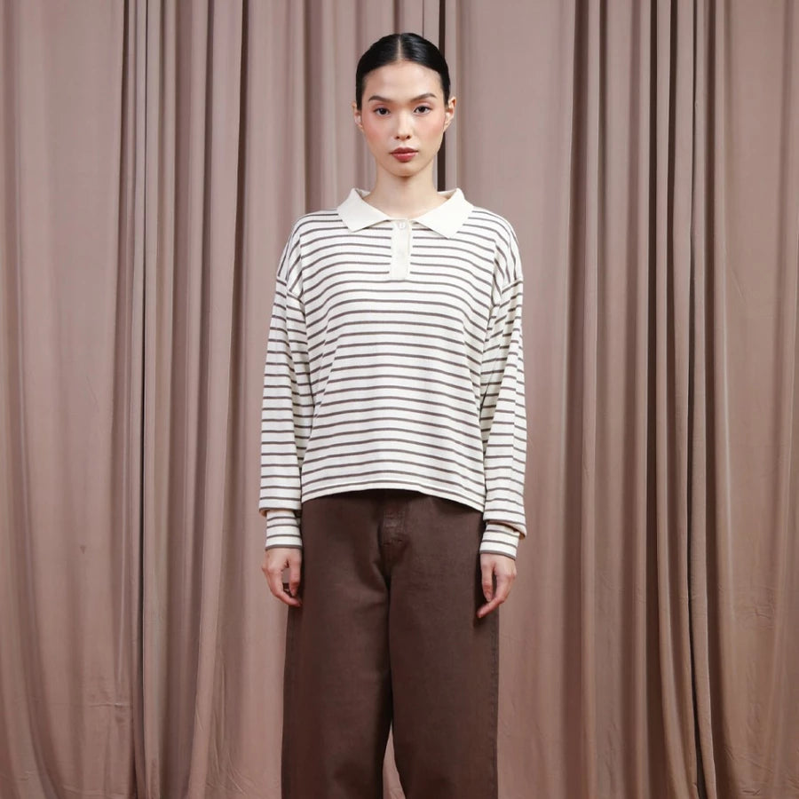 Person wearing a brown and white striped shirt by Hijabchic and brown pants against a beige curtain background