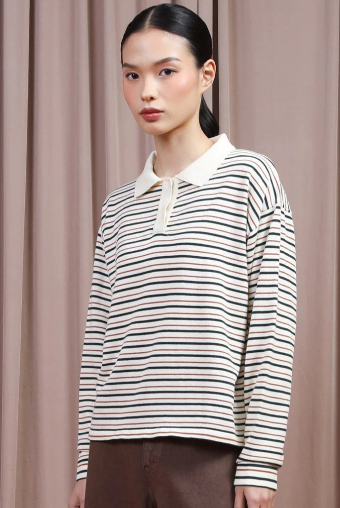 A model wearing a long-sleeve polo shirt by Hijabchic with thin horizontal green and beige stripes on an off-white base, paired with brown trousers, set against a backdrop of draped beige curtains.