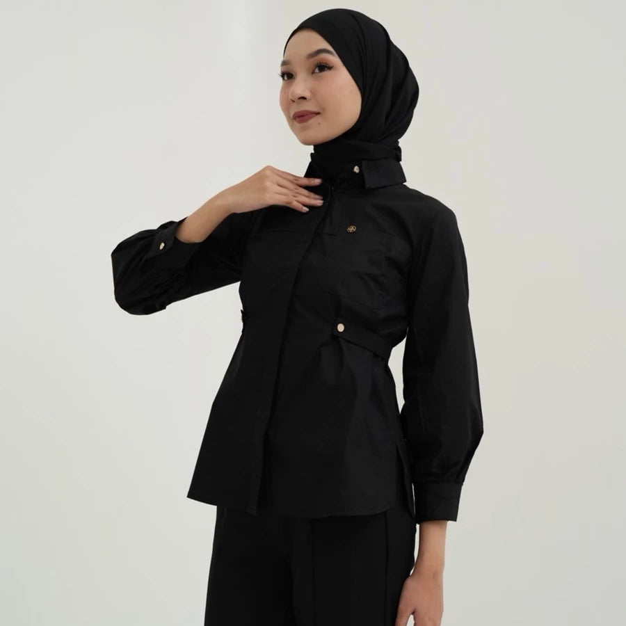 Woman wearing a black blouse by Tunique with a matching hijab against a plain background