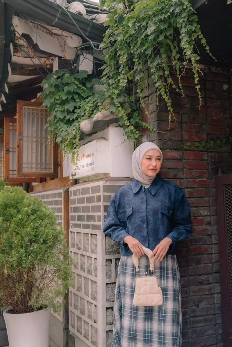 A woman wearing a blue floral long-sleeve outer by Haidee Orlin and a blue and white plaid skirt, paired with a light beige hijab and holding a small beige handbag, standing in front of a brick wall with green ivy in a courtyard setting.