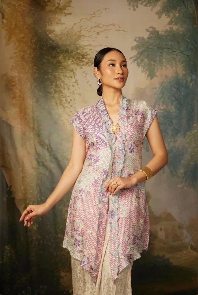 A woman wearing a lavender colored, long-sleeved velvet top by Kartinis Label with detail pita and manik, standing in front of a painted backdrop.