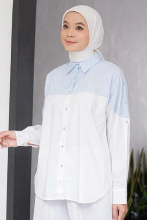 Woman wearing a light blue and white shirt by Casa Elana with a hijab against a gray wall.
