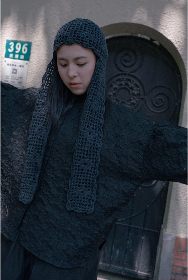 A person wearing a dark charcoal gray textured top by Yestoday, including a crocheted beanie with a long neck drape, standing in front of a building door.