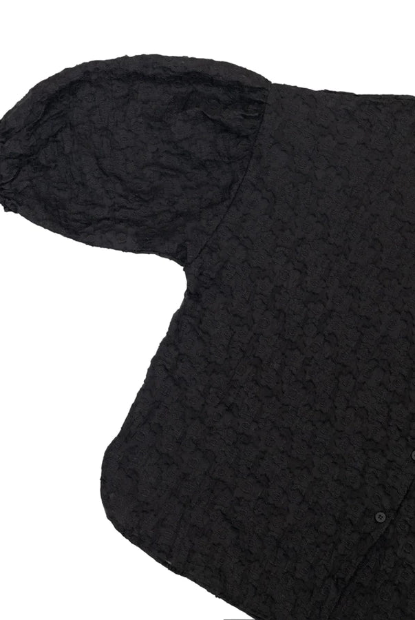 A close-up of a black textured shirt by Yestoday with a crinkled, embossed pattern, featuring a short puff sleeve and a buttoned front, shown on a plain white background.