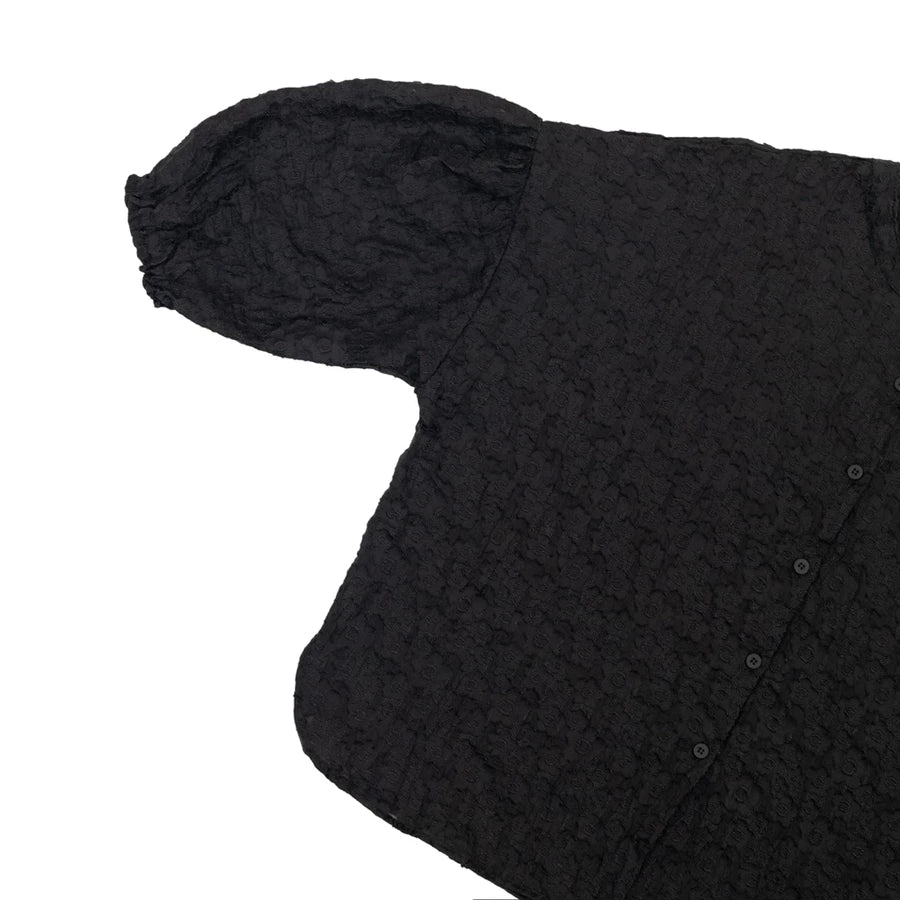 A close-up of a black textured shirt by Yestoday with a crinkled, embossed pattern, featuring a short puff sleeve and a buttoned front, shown on a plain white background.
