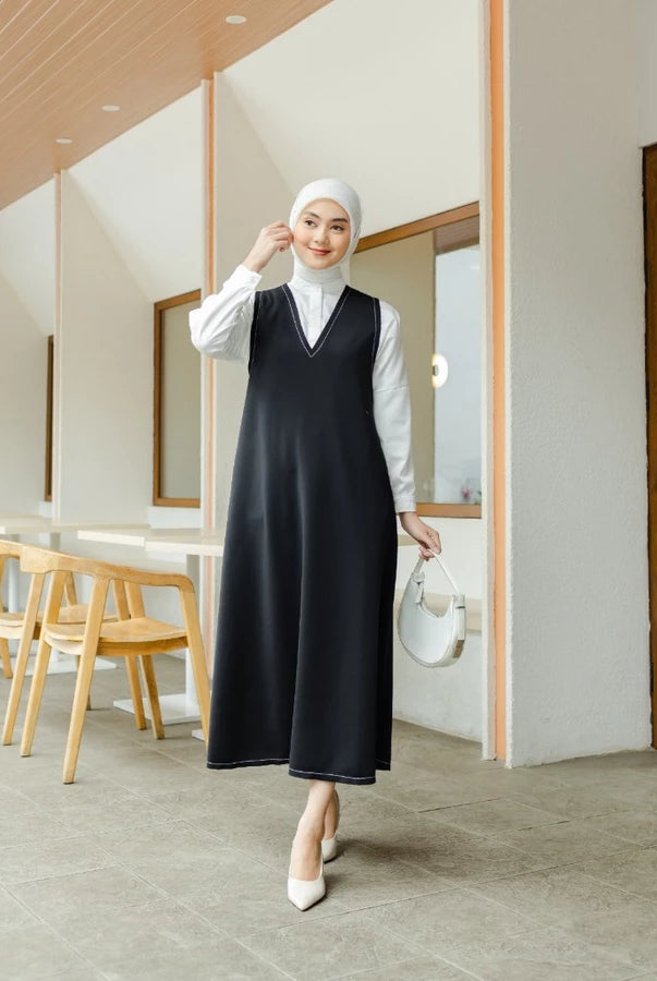Woman in a black sleeveless long dress by Abame and white hijab standing in a modern interior setting.