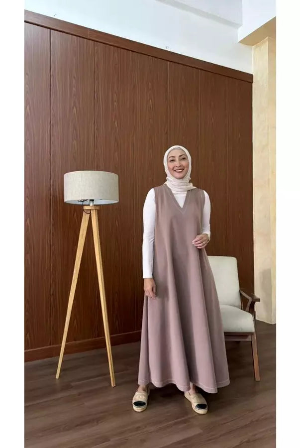 Woman in a brown long sleeveless dress by Abame standing in a room with wooden paneling and a lamp.