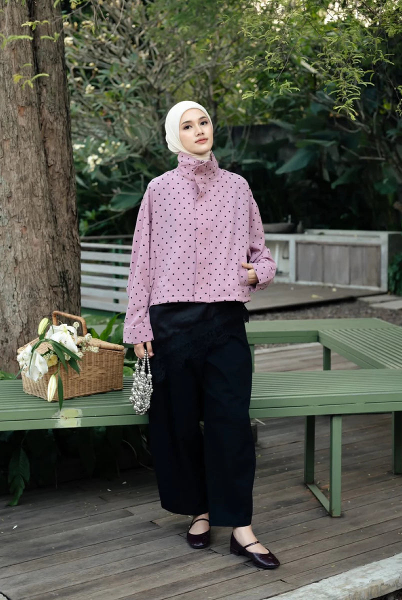 A woman wearing a light pink long-sleeve turtleneck shirt by Haidee Orlin with a small black polka dot pattern, paired with black wide-leg pants, a white hijab, and dark flat shoes, standing on a wooden deck in a garden setting.