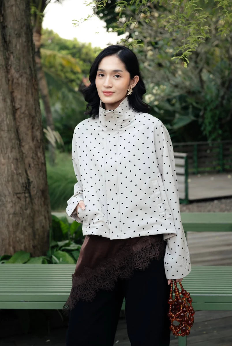 A woman standing outdoors in a park-like setting wearing a white long-sleeve mock-neck shirt by Haidee Orlin with black polka dots, a dark brown lace-trimmed wrap or skirt layer, black pants, and holding a red beaded handbag.