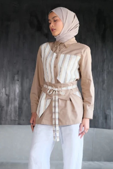 Woman wearing a beige blouse by Tunique with a striped tie and white pants against a dark textured wall.