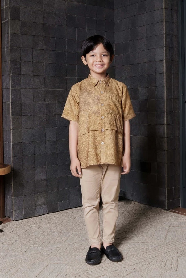 A boy wearing a patterned gold shirt by Jenna & Kaia and beige pants standing indoors against a dark wall.
