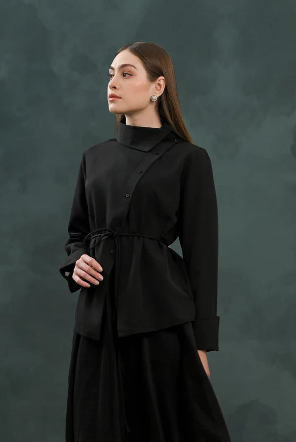 A woman modeling a black long-sleeve wrap-style blouse by Kasa Heritage with a drawstring waist, paired with a black skirt, set against a dark textured studio backdrop.