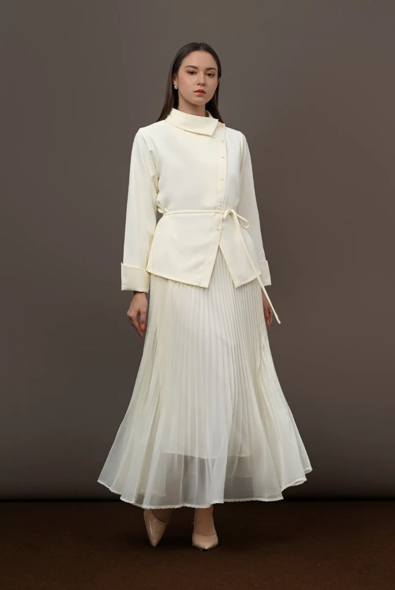 A model stands against a dark brown studio backdrop wearing a cream-colored two-piece outfit consisting of a long-sleeve wrap-style blouse by Kasa Heritage with a tie belt and a matching pleated maxi skirt.