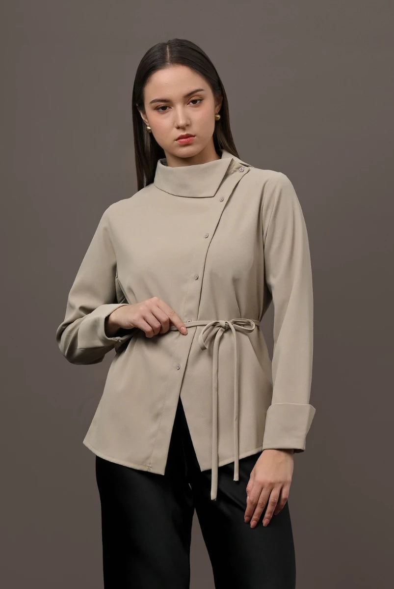 A woman modeling a beige long-sleeve wrap-style blouse by Kasa Heritage with a high mock neck and a drawstring tie at the waist, paired with black pants, against a solid dark gray studio background.