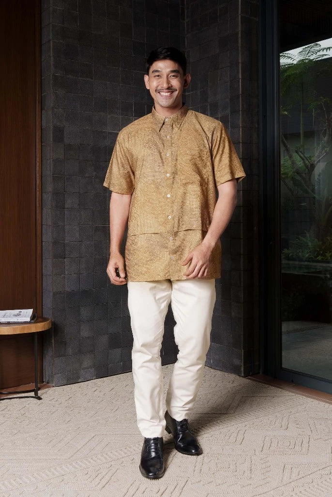 Man in a gold shirt by Jenna & Kaia and white pants standing in a modern indoor setting.