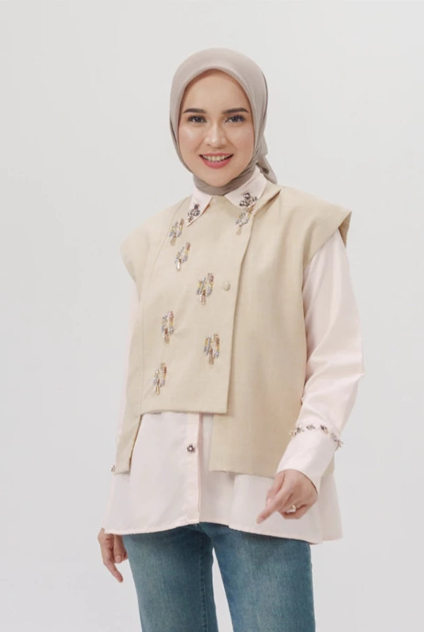 Woman wearing a khaki embroidered vest over a white shirt with a plain background