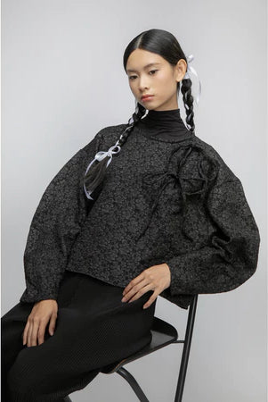 Woman wearing a black textured blouse by Yestoday with puffed sleeves and black pants, sitting on a chair against a light gray background.