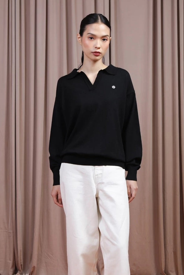 A model stands against a brown curtain backdrop wearing a black long-sleeve polo-style knit top by Hijabchic with a small white embroidered logo on the chest, paired with white pants.