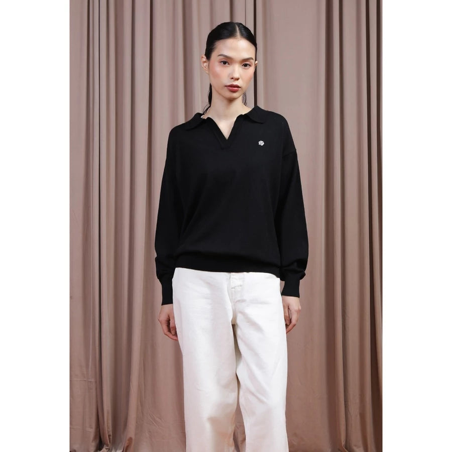 A model stands against a brown curtain backdrop wearing a black long-sleeve polo-style knit top by Hijabchic with a small white embroidered logo on the chest, paired with white pants.