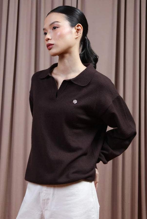 A woman wearing a dark brown long-sleeve polo shirt by Hijabchic with a small embroidered logo on the chest, paired with white pants, standing in front of a brown curtain backdrop.