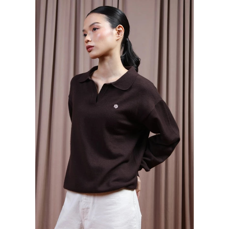 A woman wearing a dark brown long-sleeve polo shirt by Hijabchic with a small embroidered logo on the chest, paired with white pants, standing in front of a brown curtain backdrop.