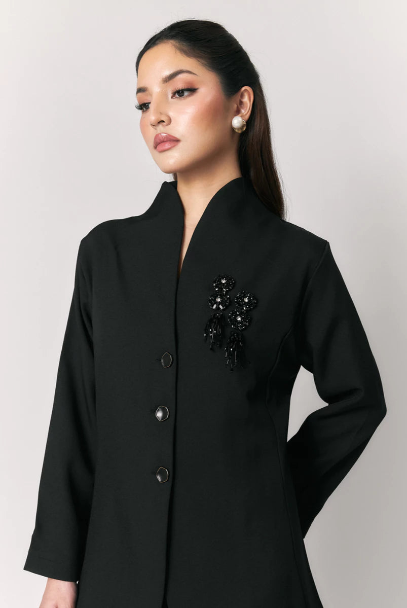 A woman wearing a black blazer by Studio Tui with a stand collar, three front buttons, and a black beaded embellishment on the left chest, photographed against a light gray studio background.