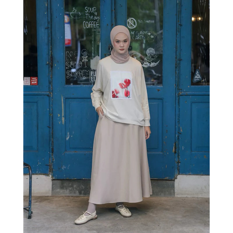 A woman wearing a beige top by Maima with a floral design on the front, paired with a long skirt and a headscarf, standing in front of a blue door.