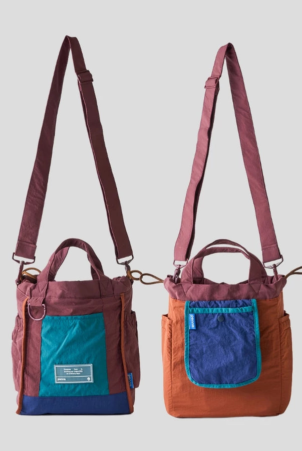 Women’s reversible tote bag by Muhajir Project featuring two looks in one design. One side comes in burgundy with navy blue accents and a teal front pocket, while the other side shows a brown body with a contrasting navy blue pocket. Designed with adjustable shoulder straps, top handles, and side drawstring details for a practical and versatile everyday bag.