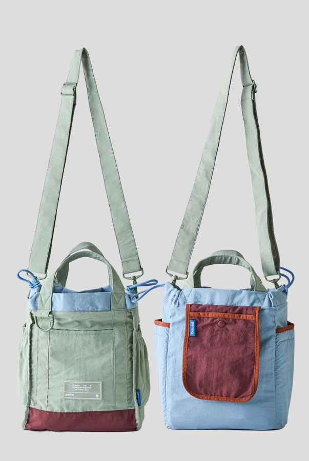 Women’s reversible tote bag by Muhajir Project featuring two looks in one design. One side comes in sage green with maroon accents and a clean front pocket, while the other side shows a light blue body with a contrasting maroon pocket. Designed with adjustable shoulder straps, top handles, and side drawstring details for a practical and versatile everyday bag.