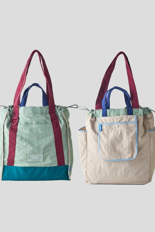Reversible tote bag by Muhajir Project displayed in two sides, showing dual-color design with maroon and light green combination on one side and grey with light blue lining pocket on the other. This versatile women’s bag features long shoulder straps, side drawstring details, and a functional front pocket, highlighting its practical and stylish two-in-one design.