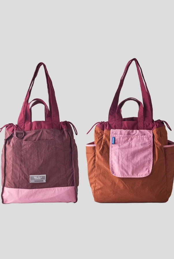 Reversible tote bag by Muhajir Project displayed in two sides, showing dual-color design with maroon and pink combination on one side and brown with pink pocket on the other. This versatile women’s bag features long shoulder straps, side drawstring details, and a functional front pocket, highlighting its practical and stylish two-in-one design.