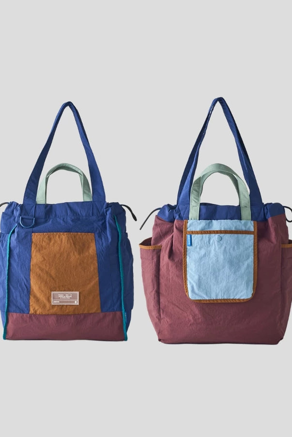 Reversible tote bag by Muhajir Project displayed in two sides, showing dual-color design with blue and brown combination on one side and maroon with light blue pocket on the other. This versatile women’s bag features long shoulder straps, side drawstring details, and a functional front pocket, highlighting its practical and stylish two-in-one design.