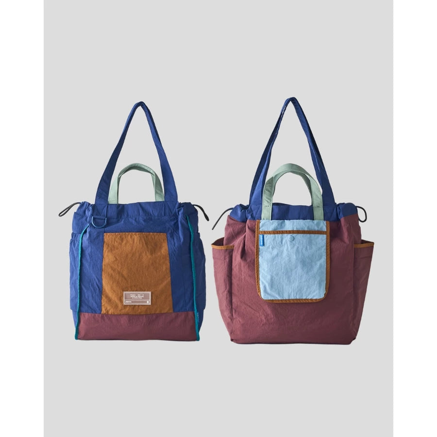 Reversible tote bag by Muhajir Project displayed in two sides, showing dual-color design with blue and brown combination on one side and maroon with light blue pocket on the other. This versatile women’s bag features long shoulder straps, side drawstring details, and a functional front pocket, highlighting its practical and stylish two-in-one design.
