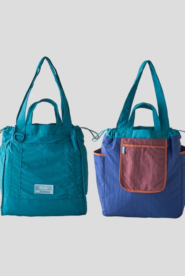 Reversible tote bag by Muhajir Project displayed in two sides, showing dual-color design teal on one side and blue with bronze pocket on the other. This versatile women’s bag features long shoulder straps, side drawstring details, and a functional front pocket, highlighting its practical and stylish two-in-one design.