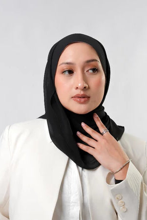 Woman wearing a black pashmina hijab by Tantri Namirah Official and white blazer against a light gray background