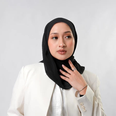 Woman wearing a black pashmina hijab by Tantri Namirah Official and white blazer against a light gray background