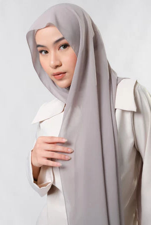 Woman wearing a light gray pashmina hijab by Tantri Namirah Official and white outfit on a plain background