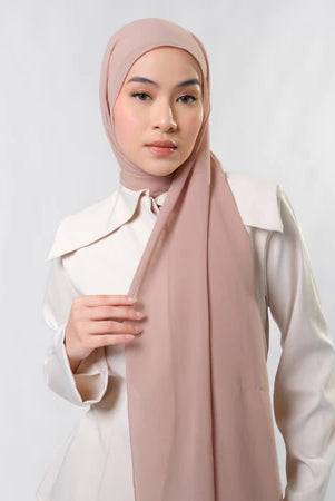 Woman wearing a light pink pashmina hijab by Tantri Namirah Official and white shirt against a plain background